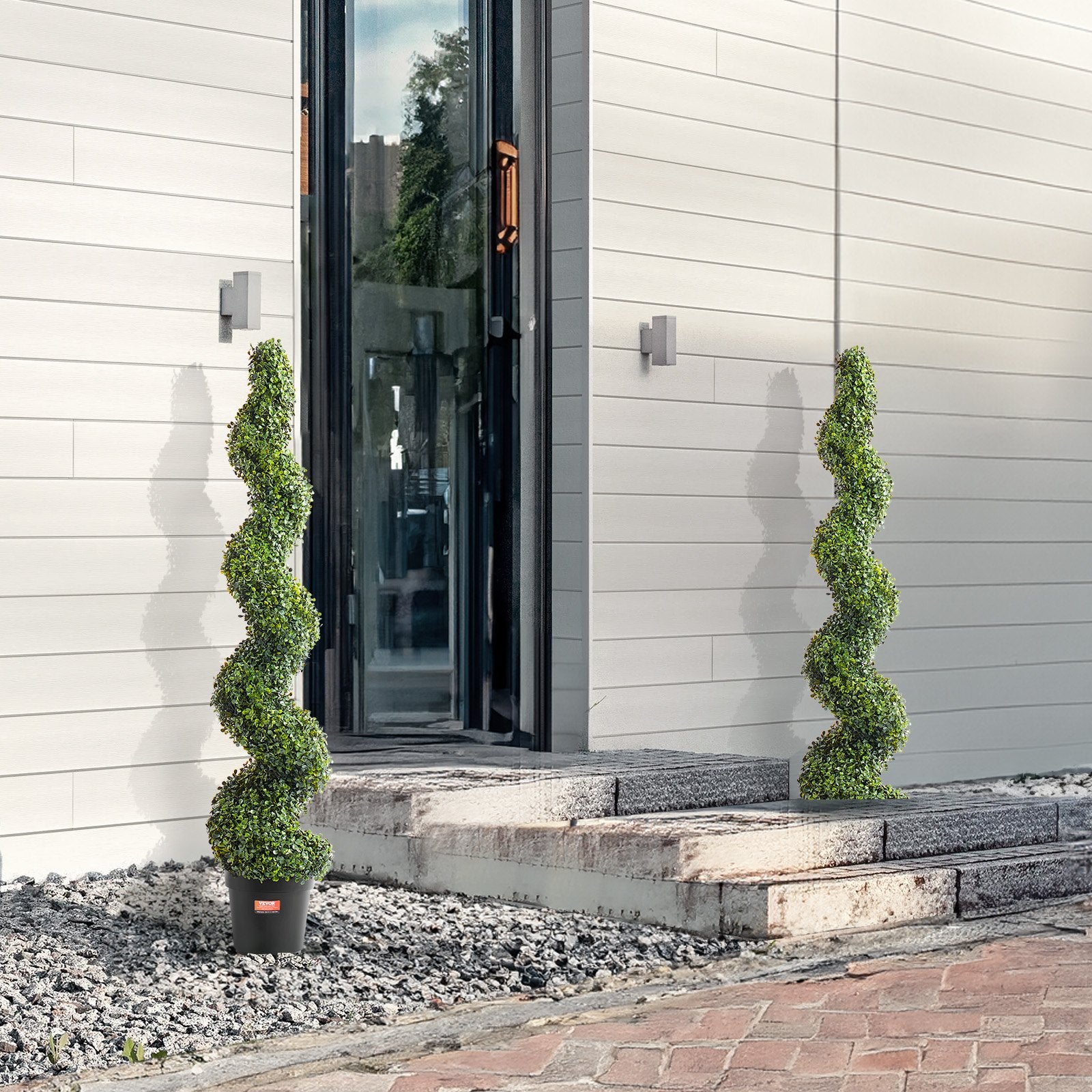 Two spiral topiary plants in pots on a stone pathway in front of a modern building.